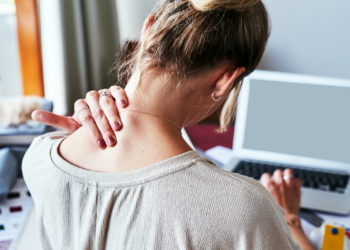 Woman seen from behind at a laptop, hand pressed to the back of her neck as she tilts her head forward.
